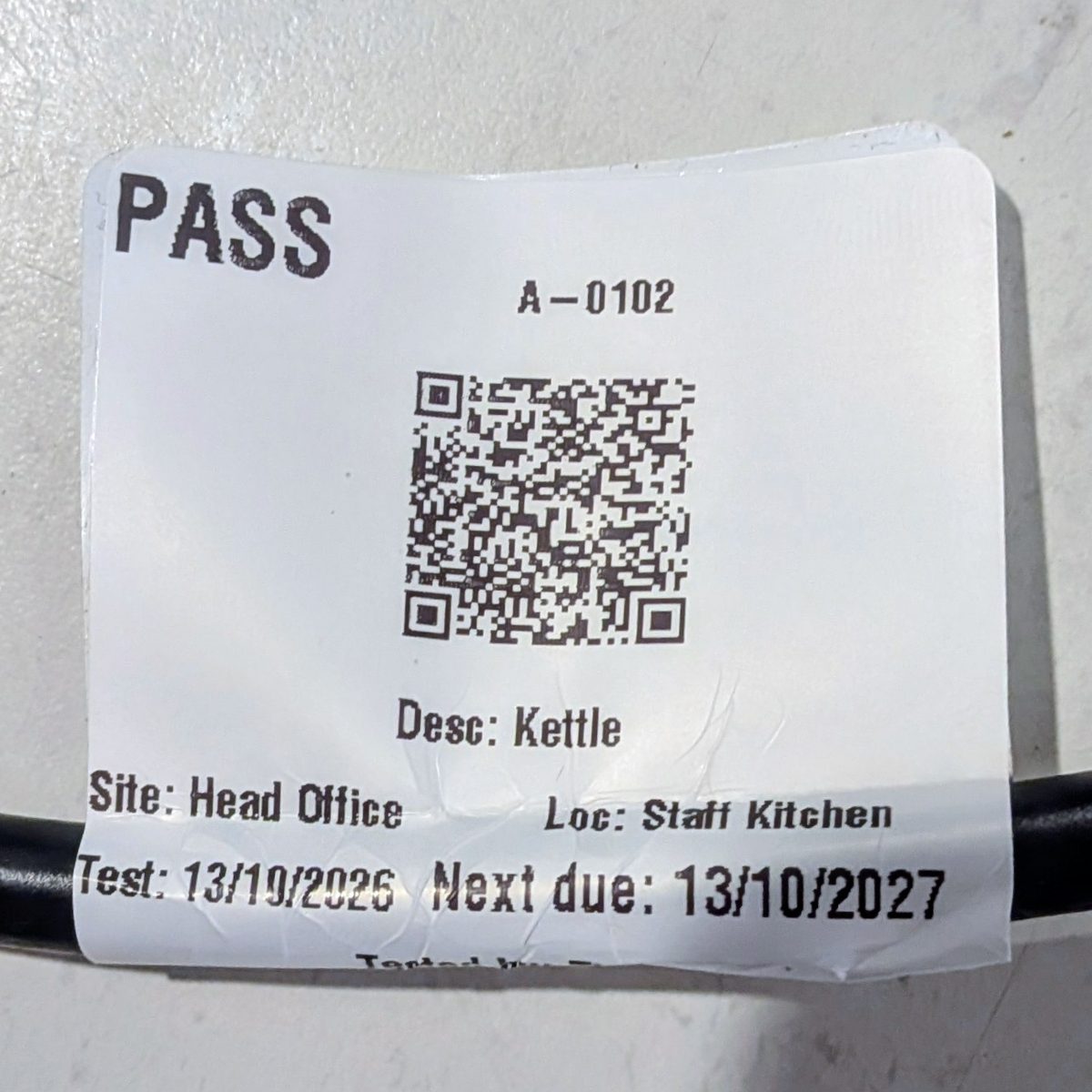 Close-up of the printed tag on the kettle lead: PASS, the appliance ID, QR code, description, site, location and dates.