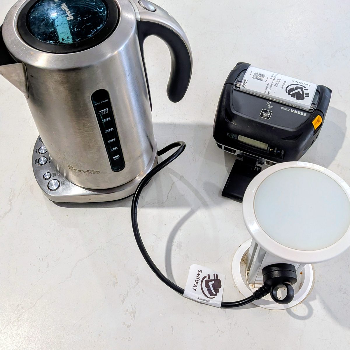 A kettle on a kitchen bench with a SwiftPAT tag on its lead and the Zebra printer beside it, the next tag coming out.